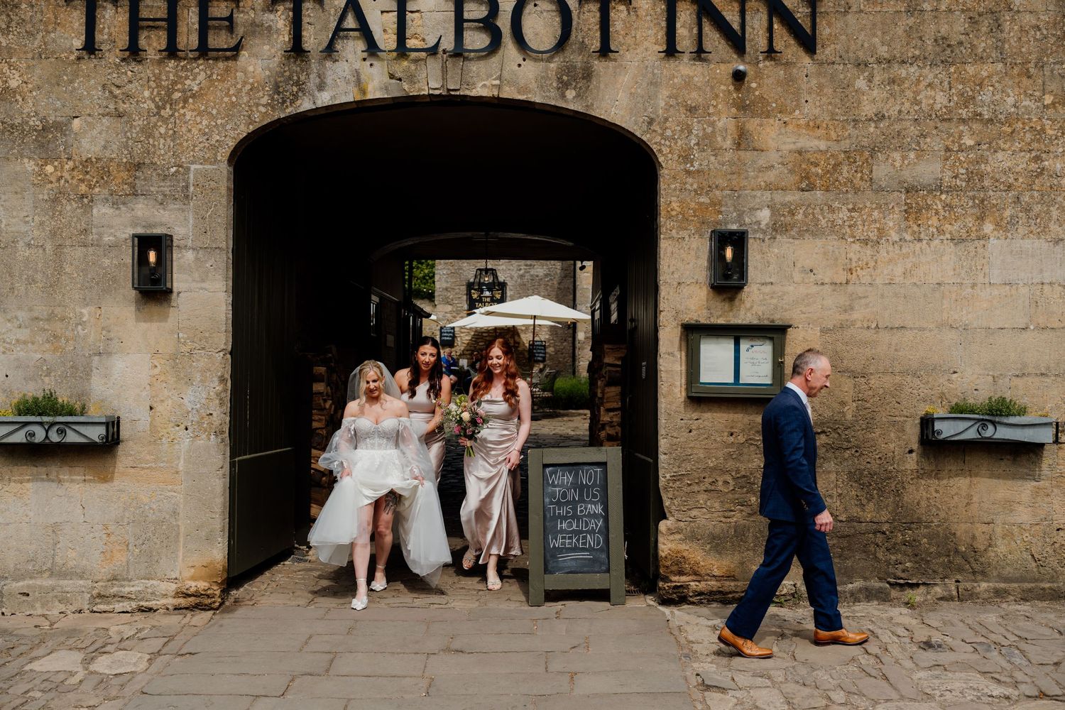 Bride and bridesmaids exiting The Talbot Inn through stone archway, with a man in blue suit walking past.