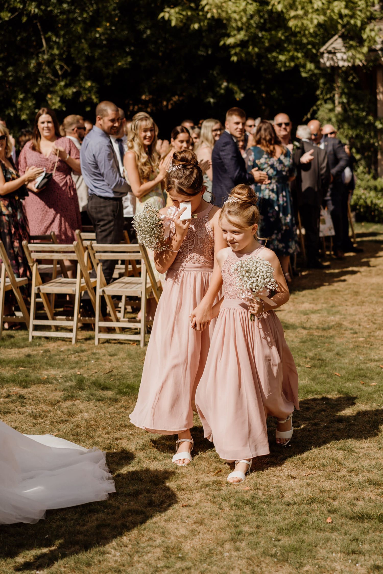 Young flower girls in blush pink dresses walk down the aisle at an outdoor garden ceremony.