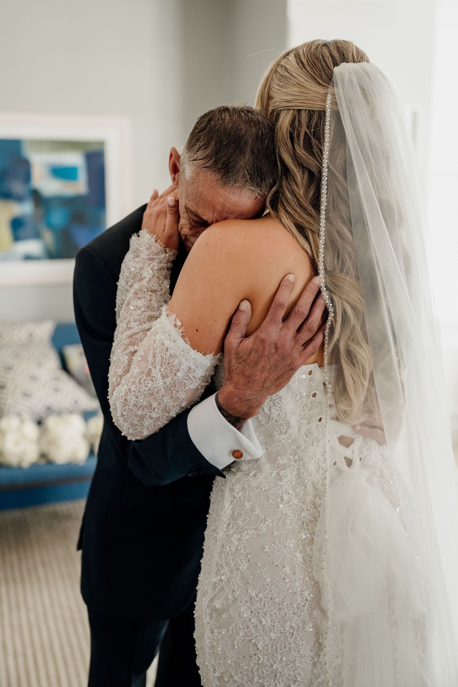 Emotional father-daughter wedding embrace, bride in beaded lace gown with veil hugging tearful dad in navy suit.