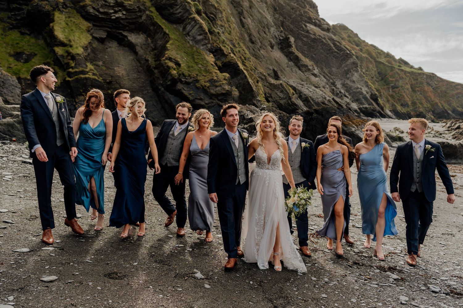Wedding party walks along a rocky beach coastline with dramatic cliffs in the background.