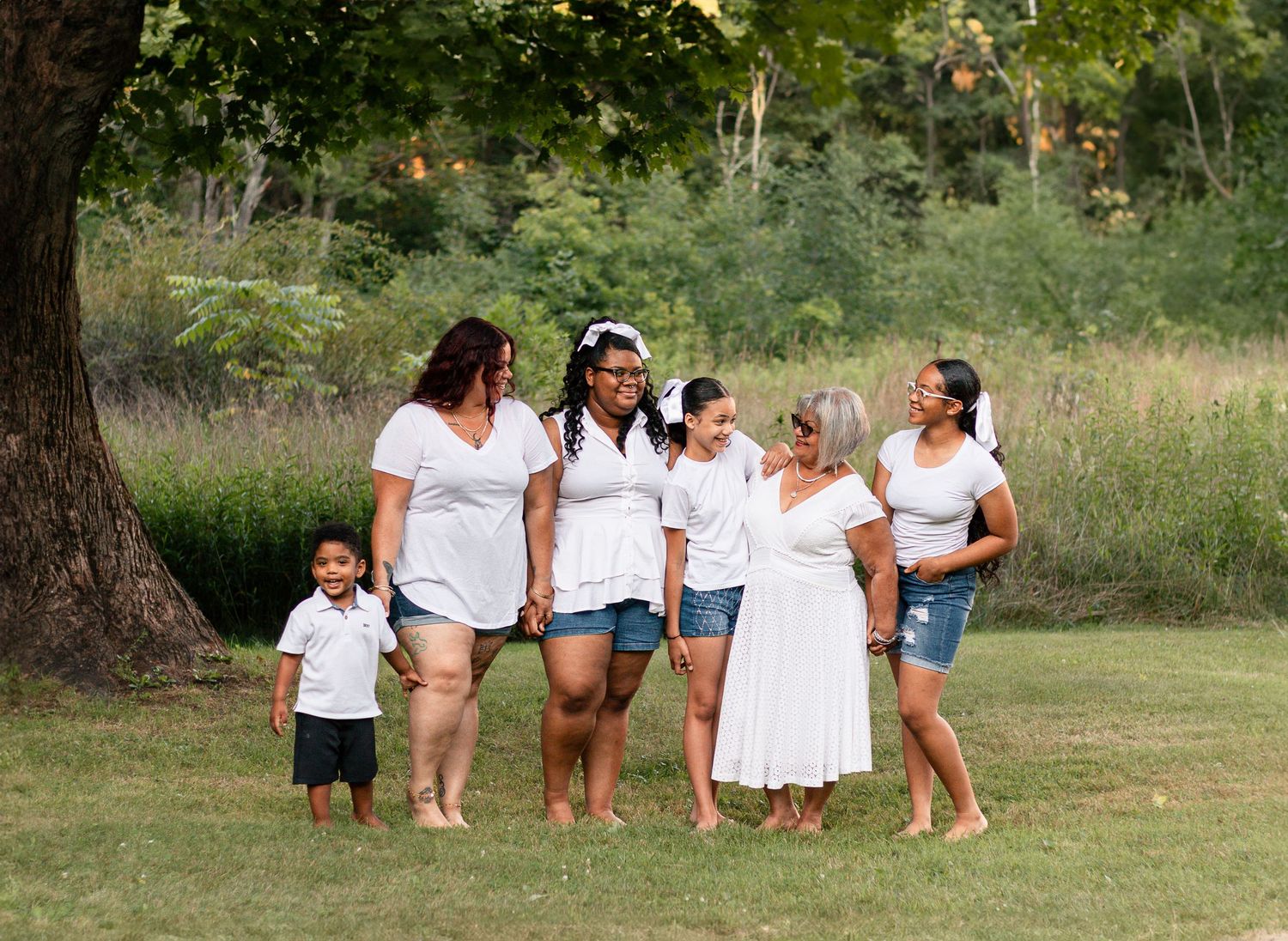 A group wearing coordinating white outfits smiles and laughs together during a casual outdoor photoshoot in a grassy field.