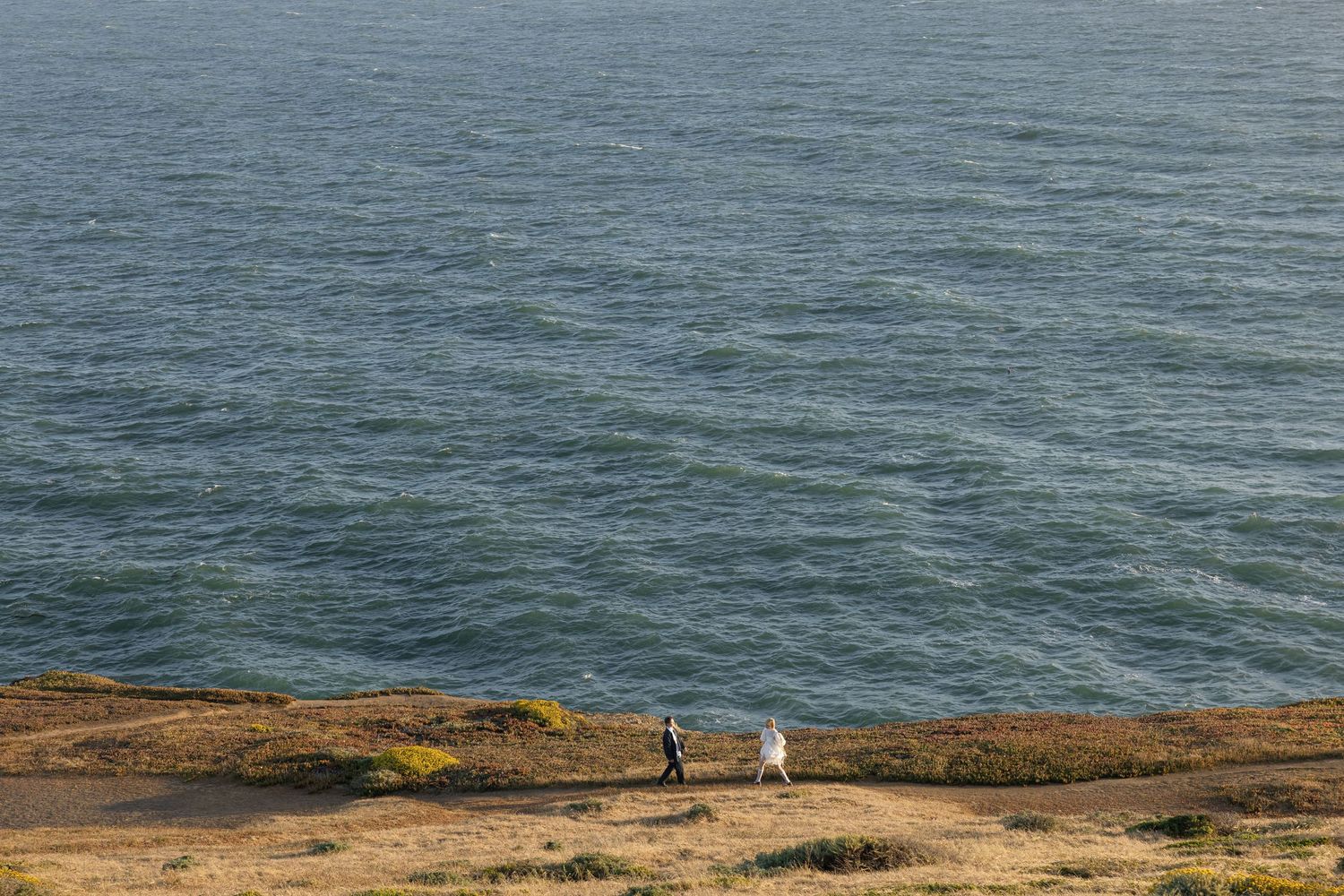 Two people walk along a rugged coastal cliff edge above the vast gray ocean waters.
