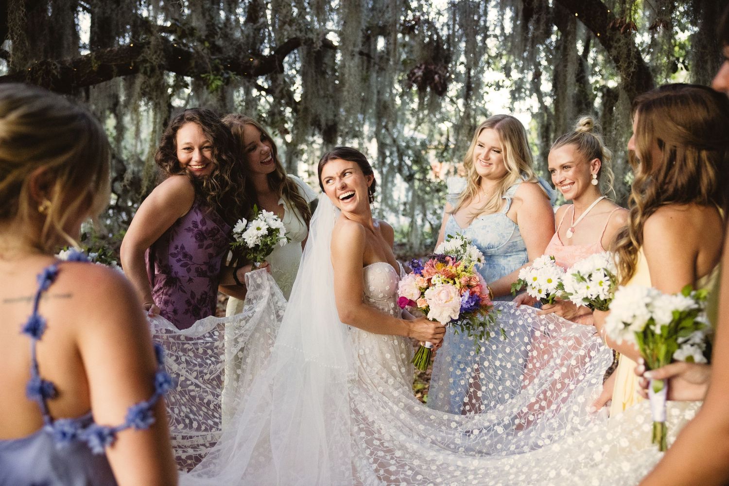 Bridesmaids in purple and floral dresses walk together under moss-draped trees during an outdoor wedding ceremony.