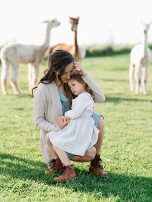 A woman holding a little girl with alpacas in the background.