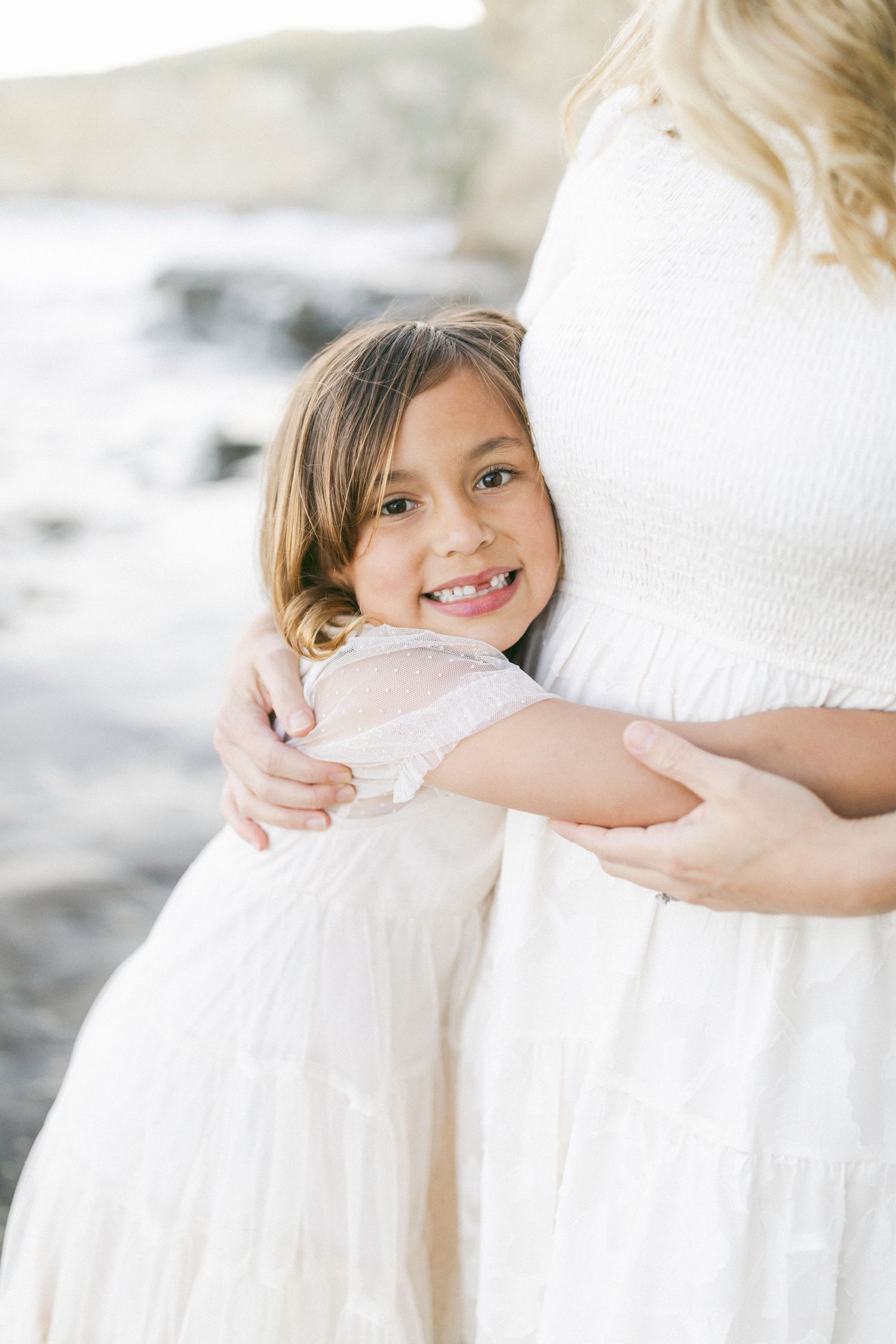 A young girl in a white dress hugs someone wearing white in a bright, soft-focus portrait setting captured by Chelsee Rawe Photography.