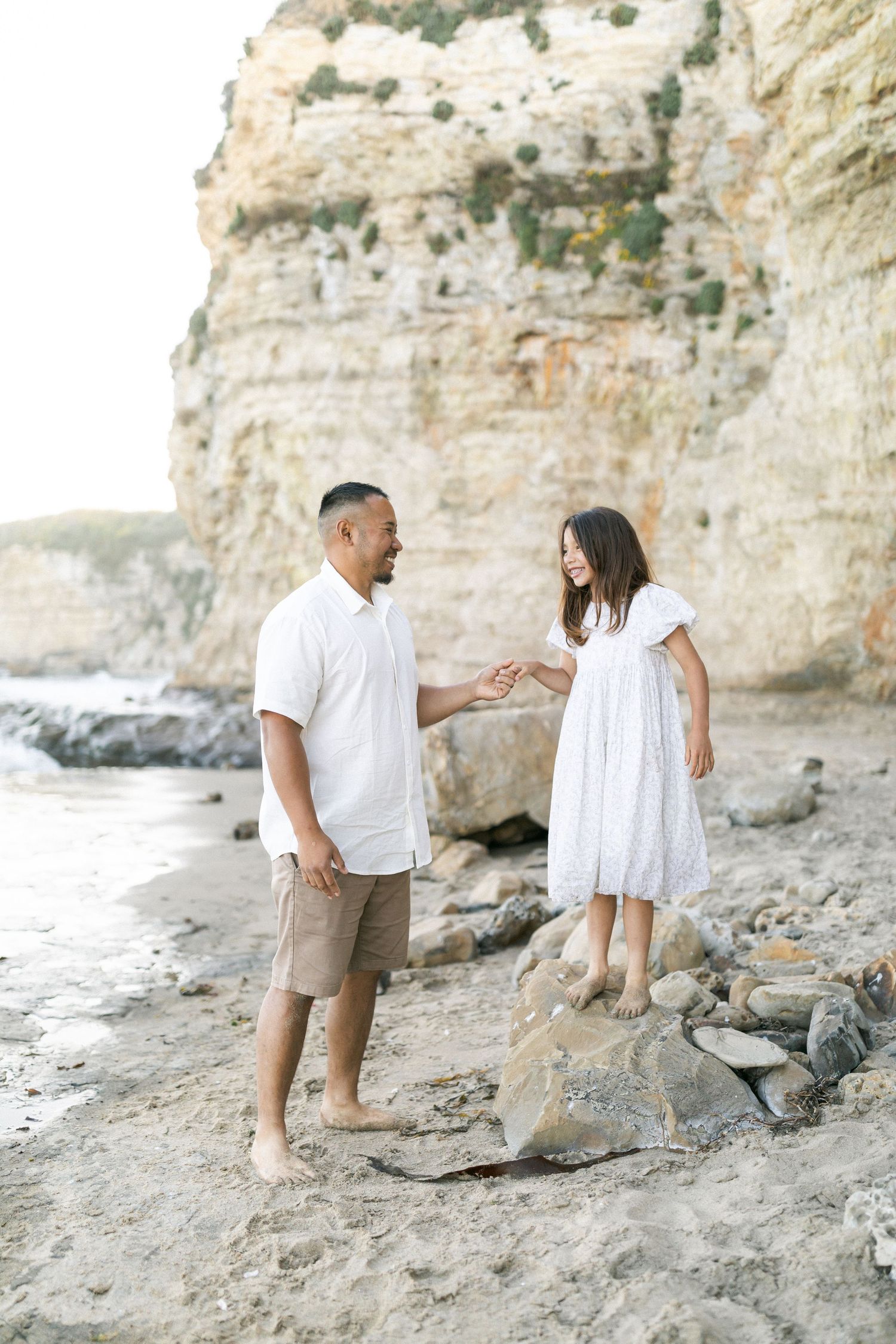 Two people in white outfits share a tender moment near coastal cliffs and rocky beach at sunset film by Chelsee Rawe .
