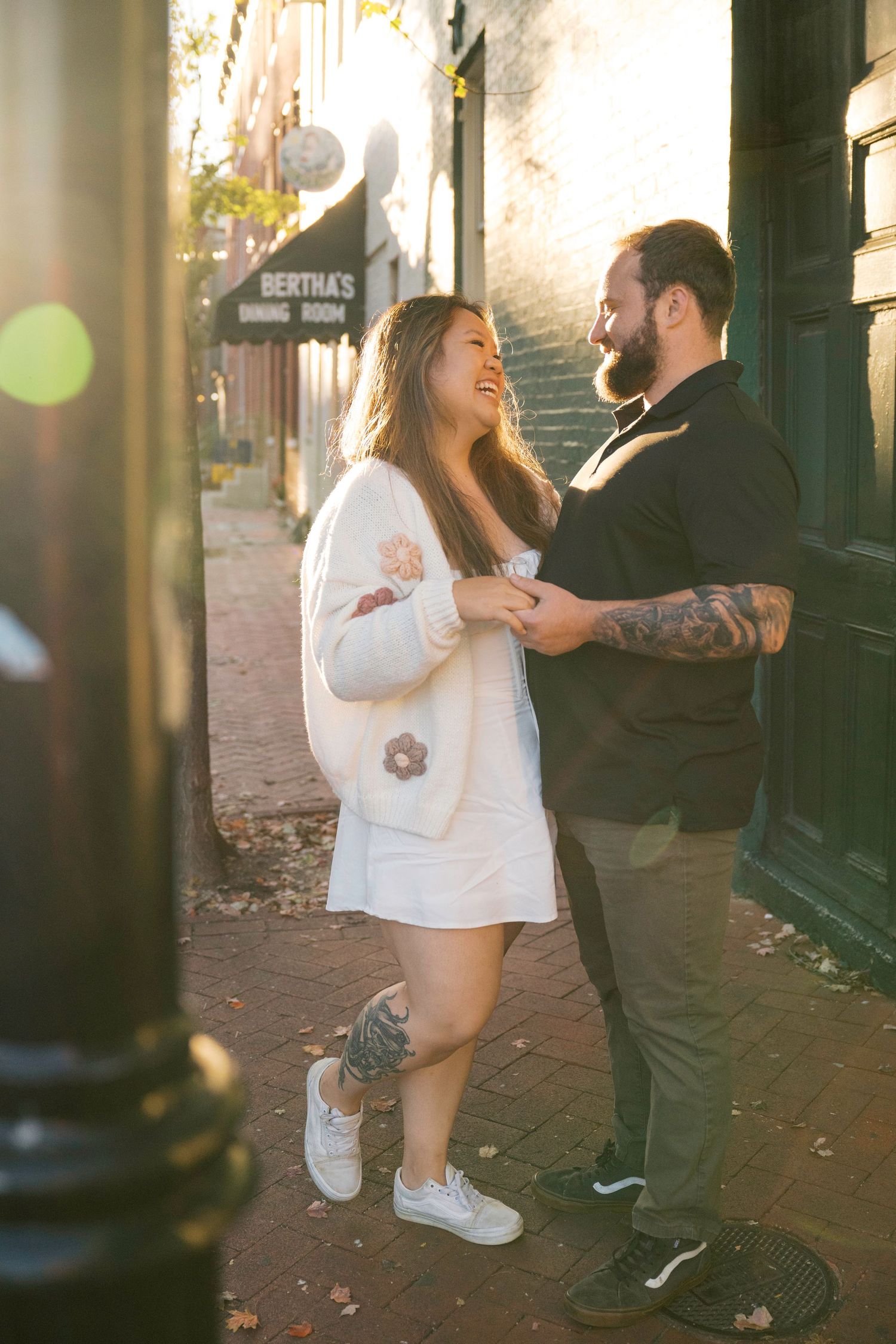 A couple shares a playful sidewalk dance moment on a sunlit city street with brick buildings and fall leaves.