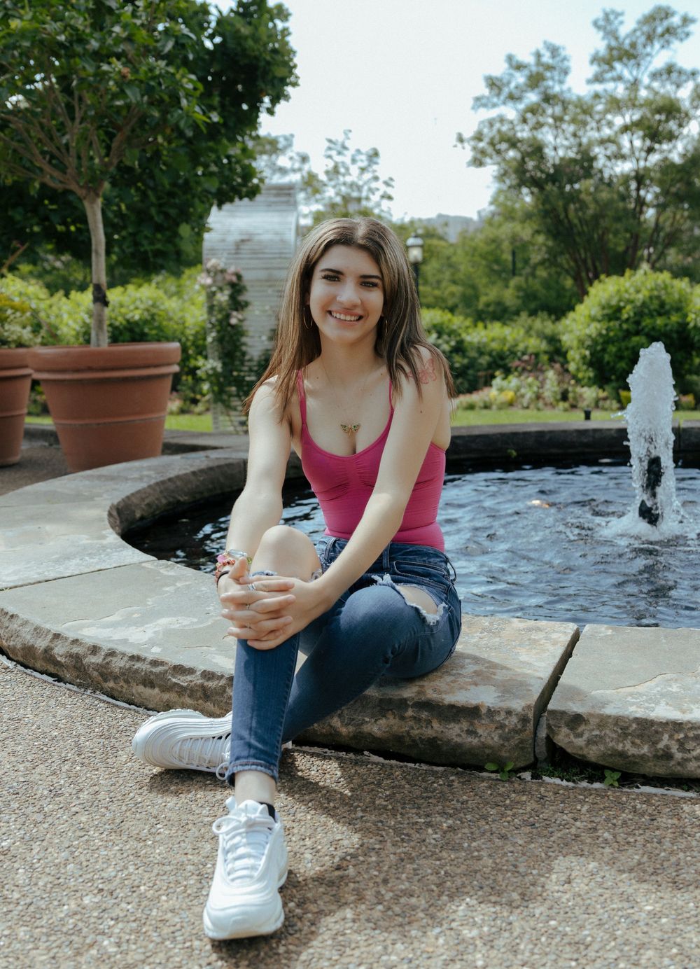 Someone in casual attire sits on the edge of a fountain in a garden setting.