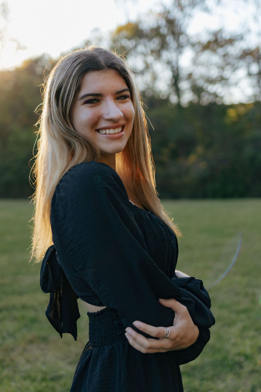 Someone wearing a black sweater smiles while embracing themselves outdoors at sunset.