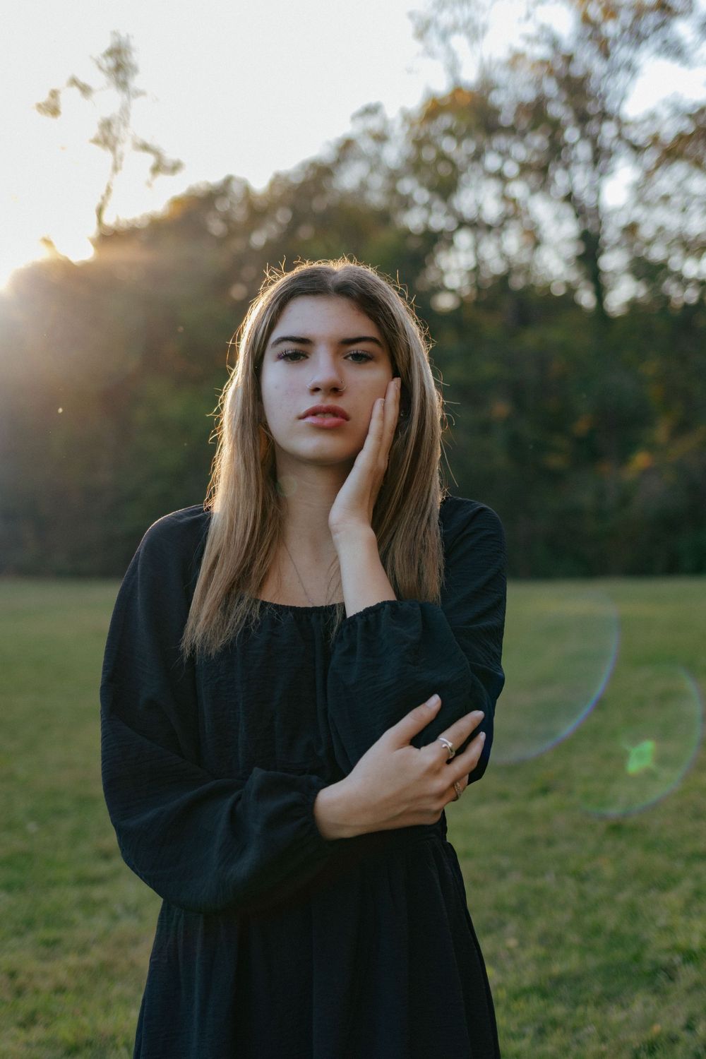 Someone in a black long-sleeve dress poses thoughtfully in an autumn field at dusk.