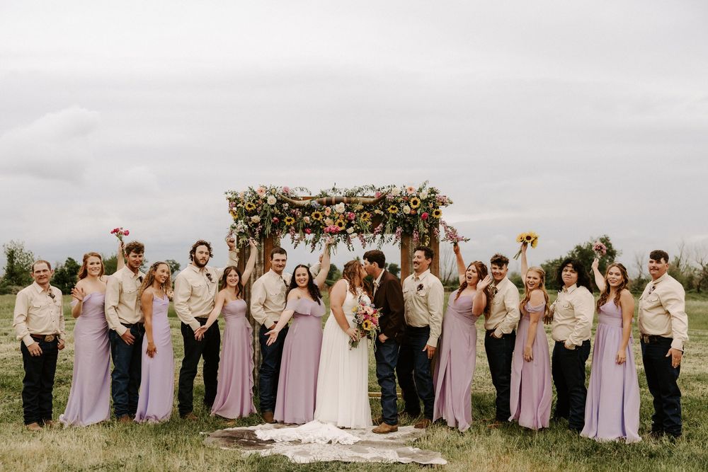 A wedding party in pastel pink and purple dresses poses together outdoors with a floral arch backdrop.