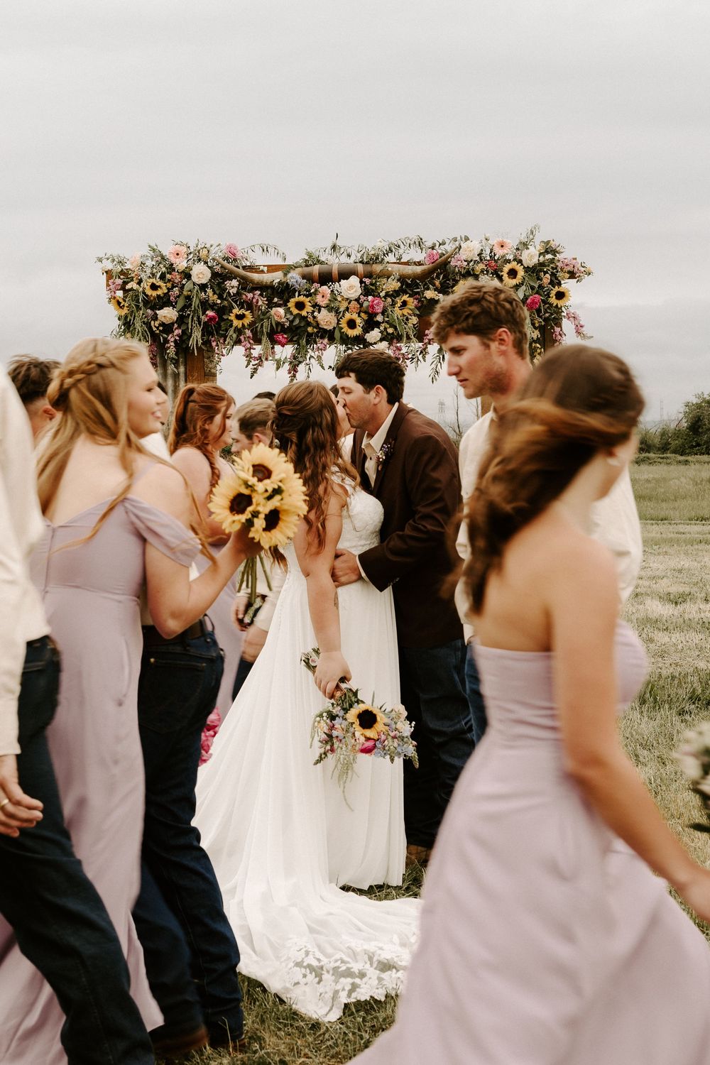 A wedding party in lavender dresses and black suits gathers outdoors with floral decorations and sunflowers.