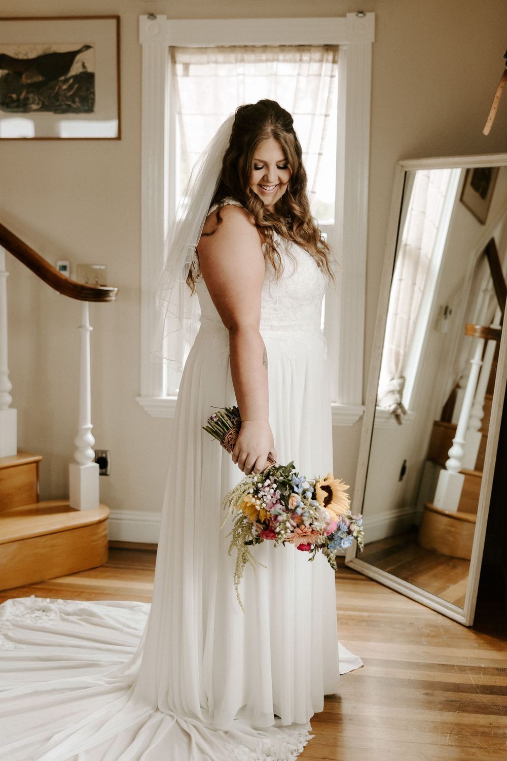 A bride in a flowing white wedding gown stands in a sunlit room with hardwood floors holding a colorful bouquet.