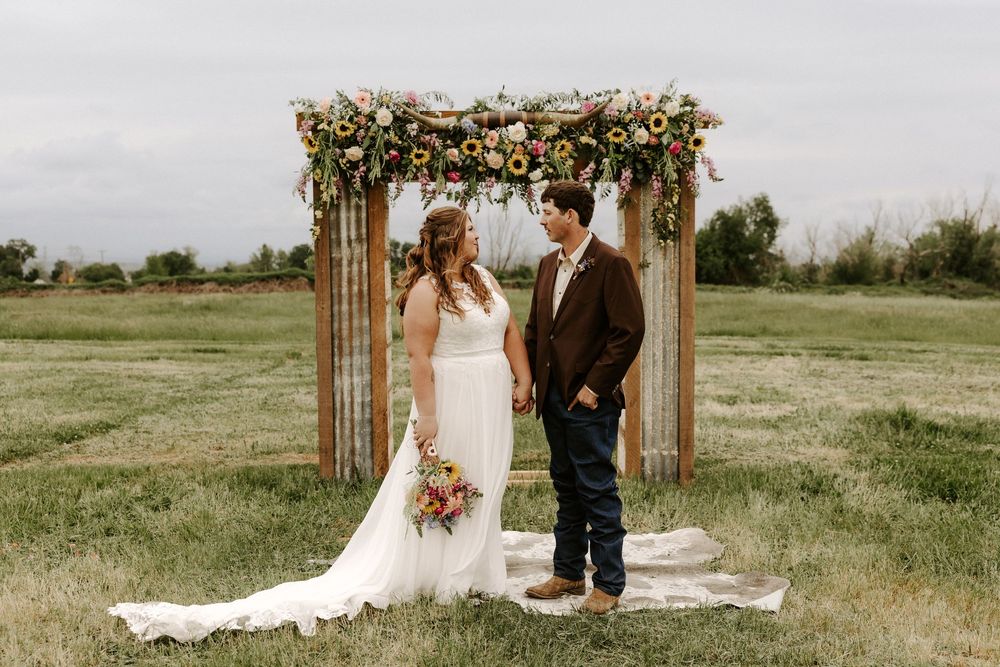 A romantic outdoor wedding ceremony takes place under a wooden arch decorated with flowers in a rustic field setting.