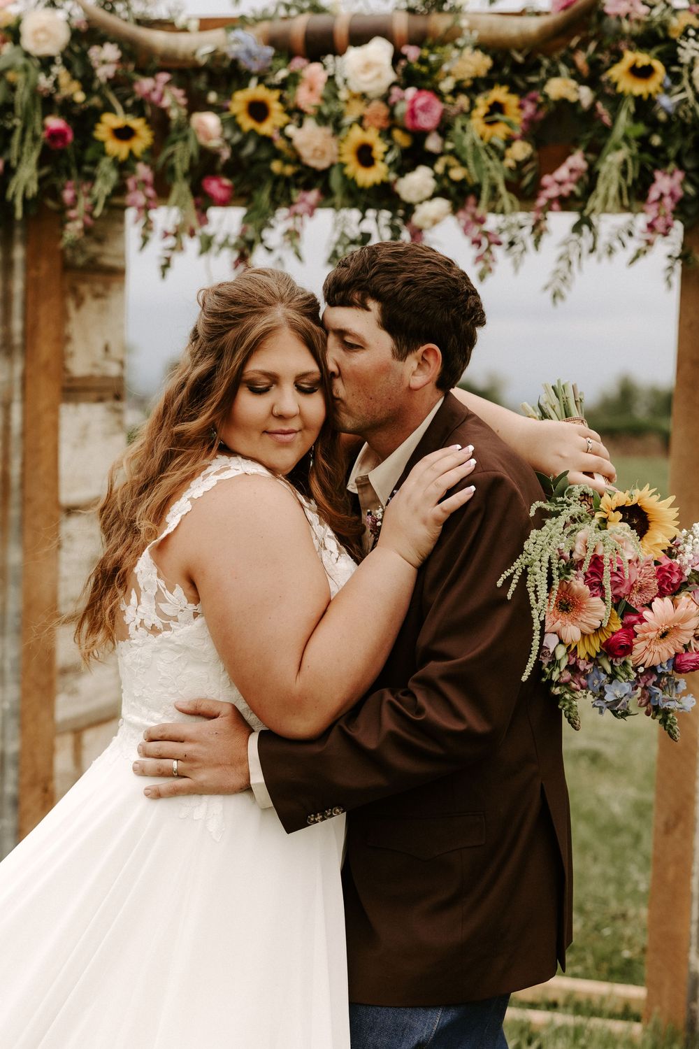Romantic wedding couple embraces under floral arch decorated with sunflowers and pink blooms during outdoor ceremony.
