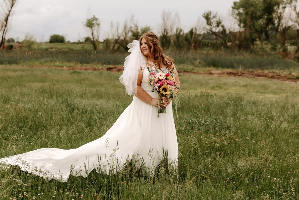 A bride in a flowing white lace dress walks through a serene meadow holding a vibrant bouquet of flowers.