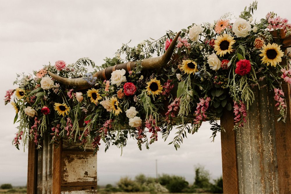 A rustic wooden arch decorated with vibrant sunflowers, roses, and greenery for an outdoor wedding ceremony.