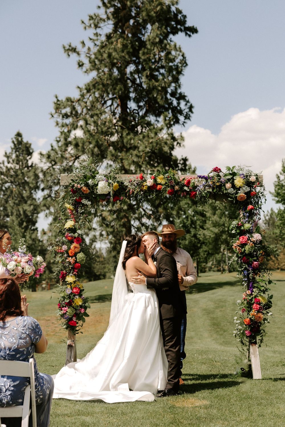 A wedding ceremony takes place under a floral arch decorated with pink and orange roses in an outdoor garden setting.