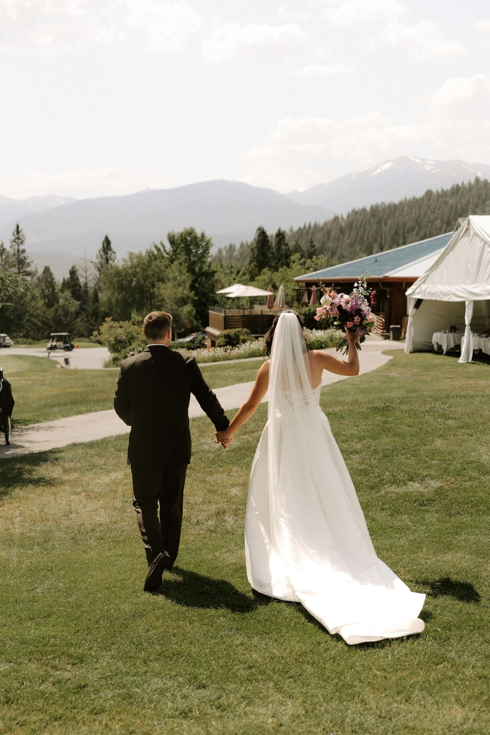 A couple walks hand in hand across a mountain venue lawn with tents and evergreen trees in the background.