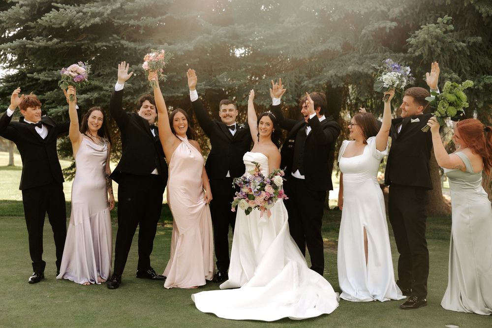 Wedding party in formal attire poses together outdoors on a sunny day in a garden setting.