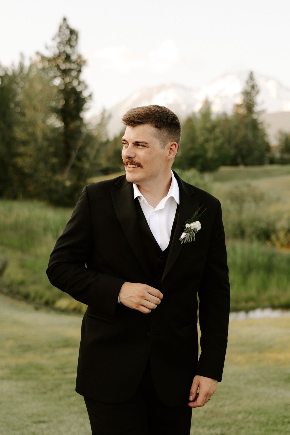 A groom in a classic black tuxedo and white boutonniere stands in front of a mountain landscape.