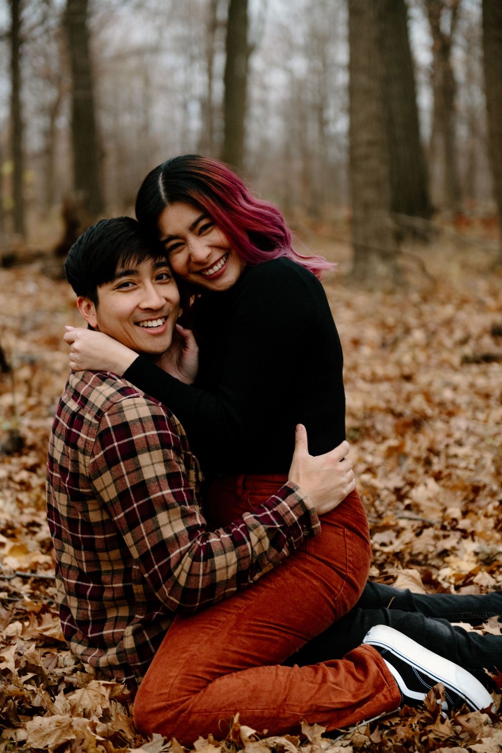 A couple lying in autumn leaves sharing a joyful embrace in a wooded area during fall.