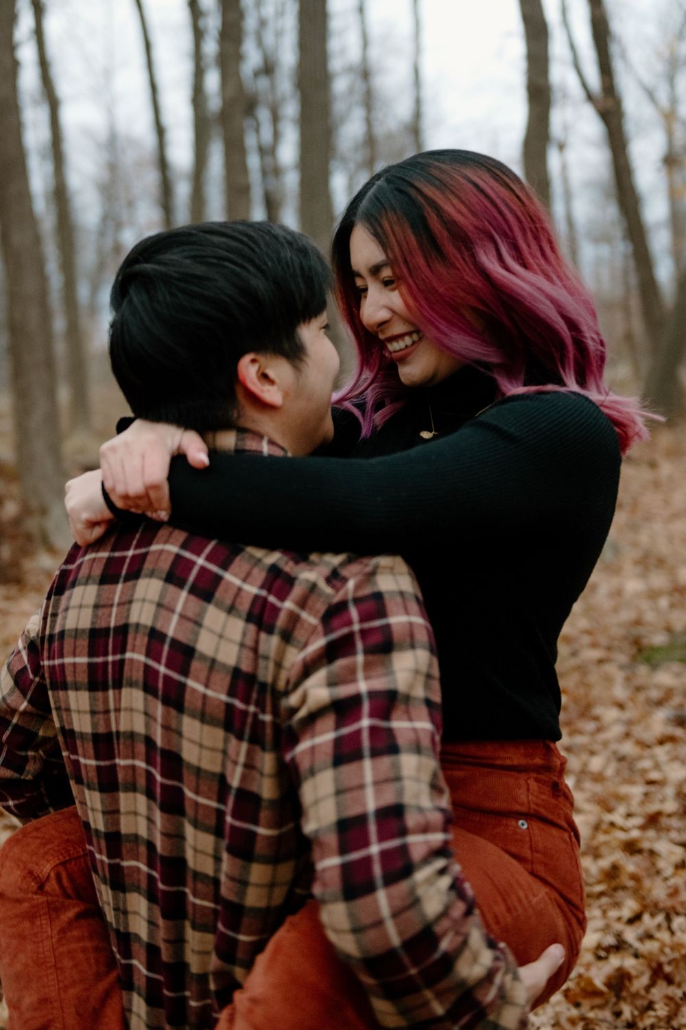 A couple embraces joyfully in autumn woods with fallen leaves, wearing plaid and black clothing.