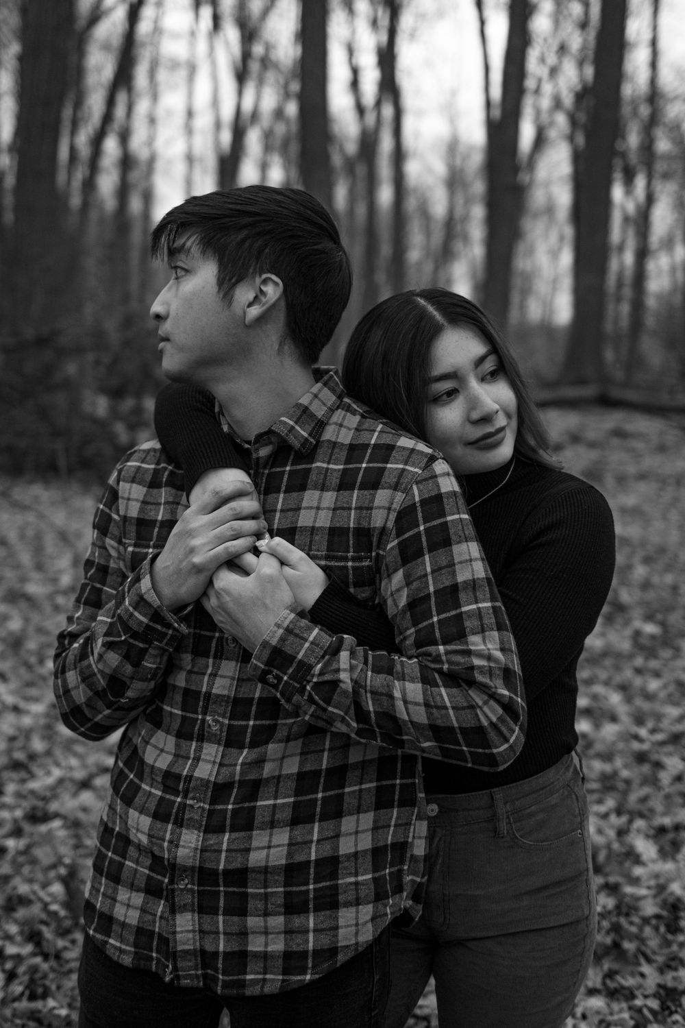 A couple embraces in a moody black and white forest setting with fallen leaves on the ground.