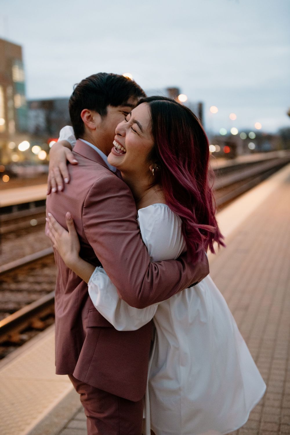 A romantic couple embraces on a train platform at sunset with city lights glowing in the background. Surprise Proposal