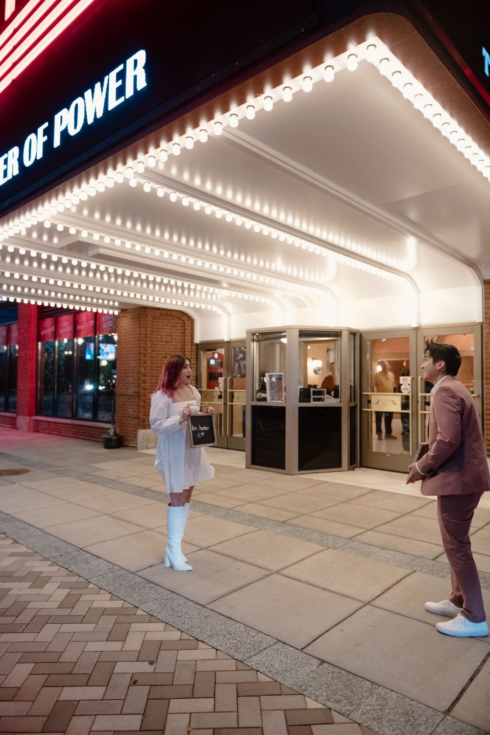 Two people stand outside a theater with 'Tower of Power' signage and bright marquee lighting.