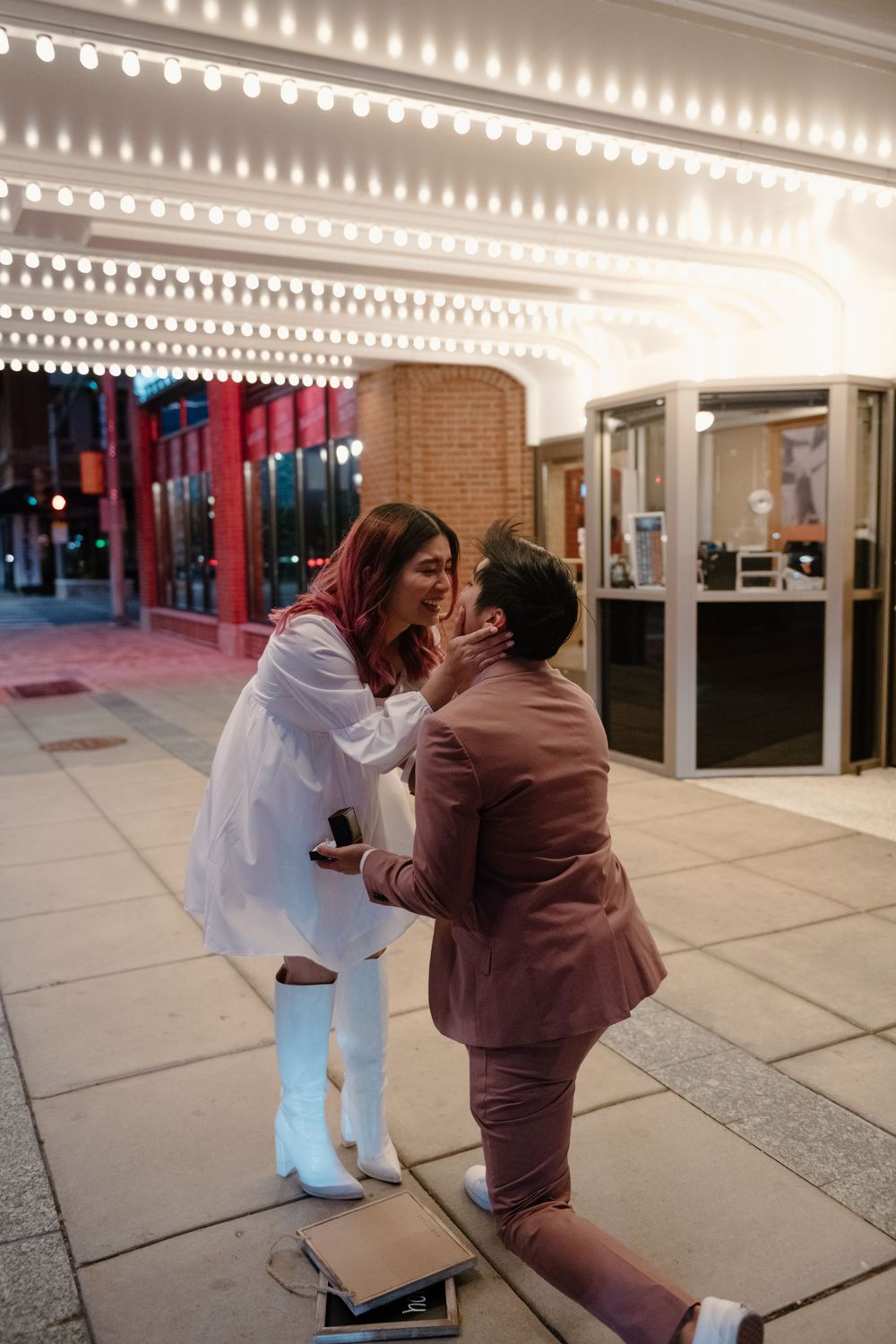 A couple embraces in a warmly lit hallway with white columns and brick walls.