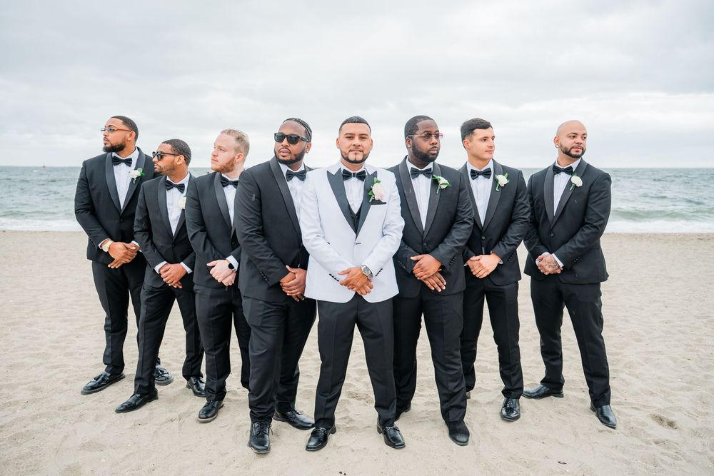 Groomsmen in black suits sitting casually on a beach showing off their matching dress shoes.
