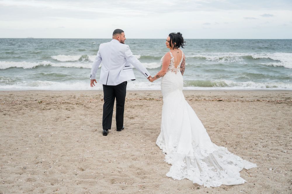 Couple holds hands while walking on beach, bride's lace dress train trailing in sand.