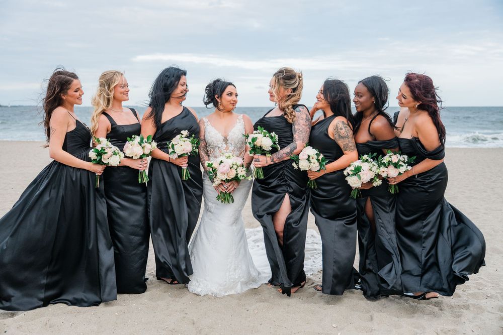 Bridesmaids in elegant black dresses pose together with the bride on a sandy beach.