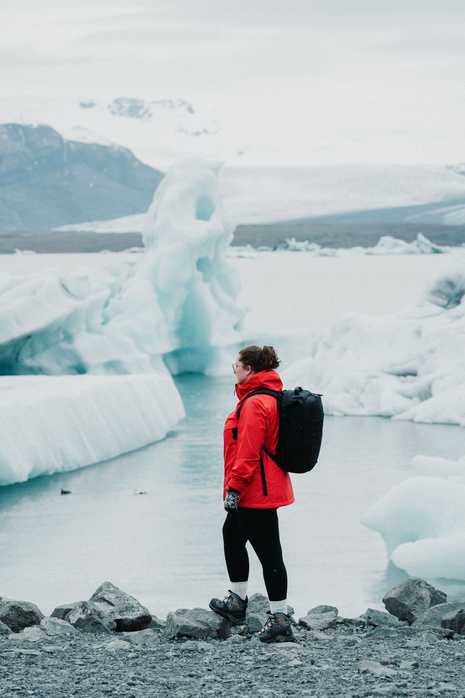 Hiker in red jacket with backpack standing among icebergs at Jokulsarlon glacier lagoon in Iceland.