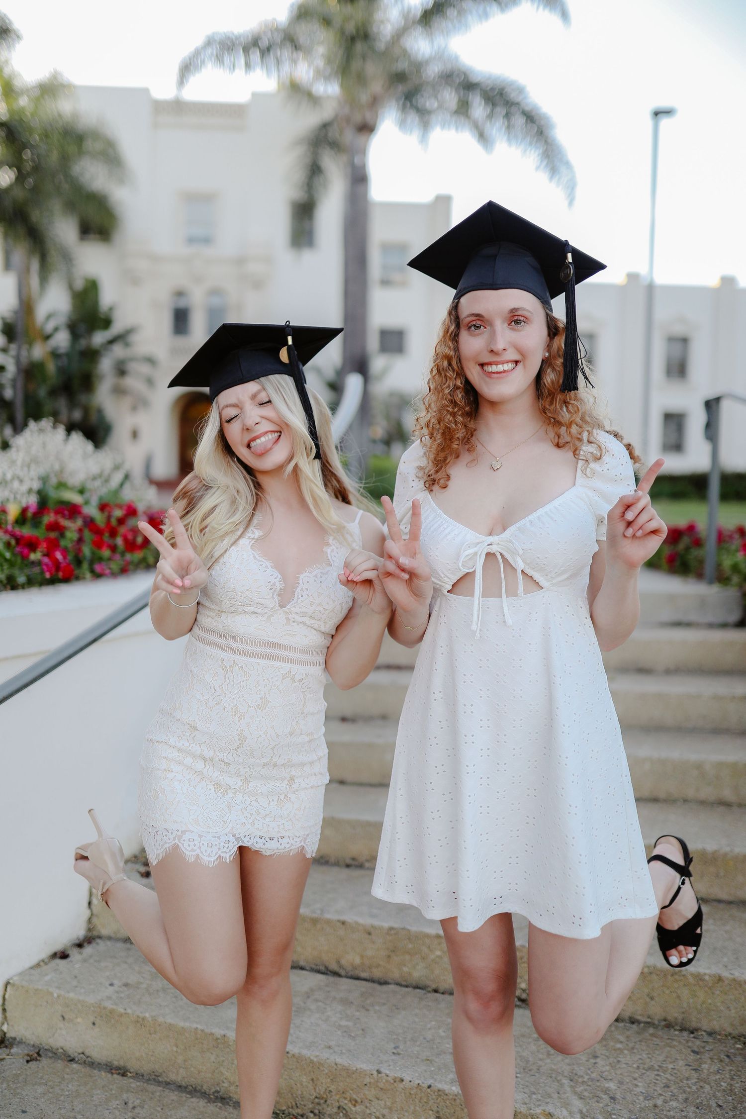 Two besties in matching white dresses strike playful poses at their campus for group graduation photos.