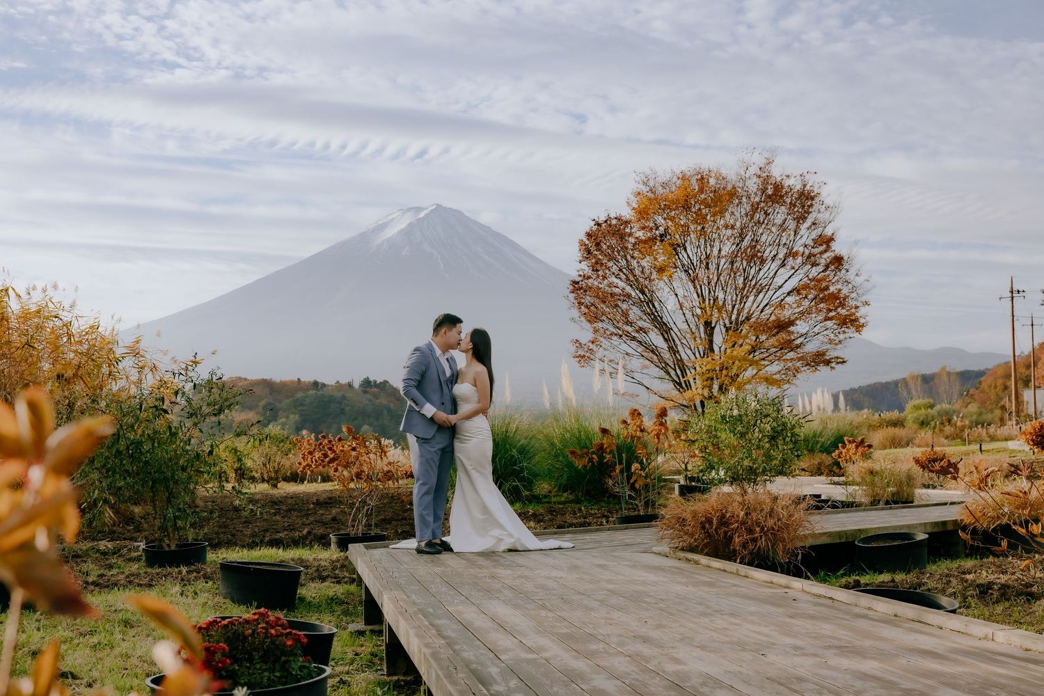 A couple embraces on a wooden boardwalk with a mountain landscape and autumn foliage in the background.