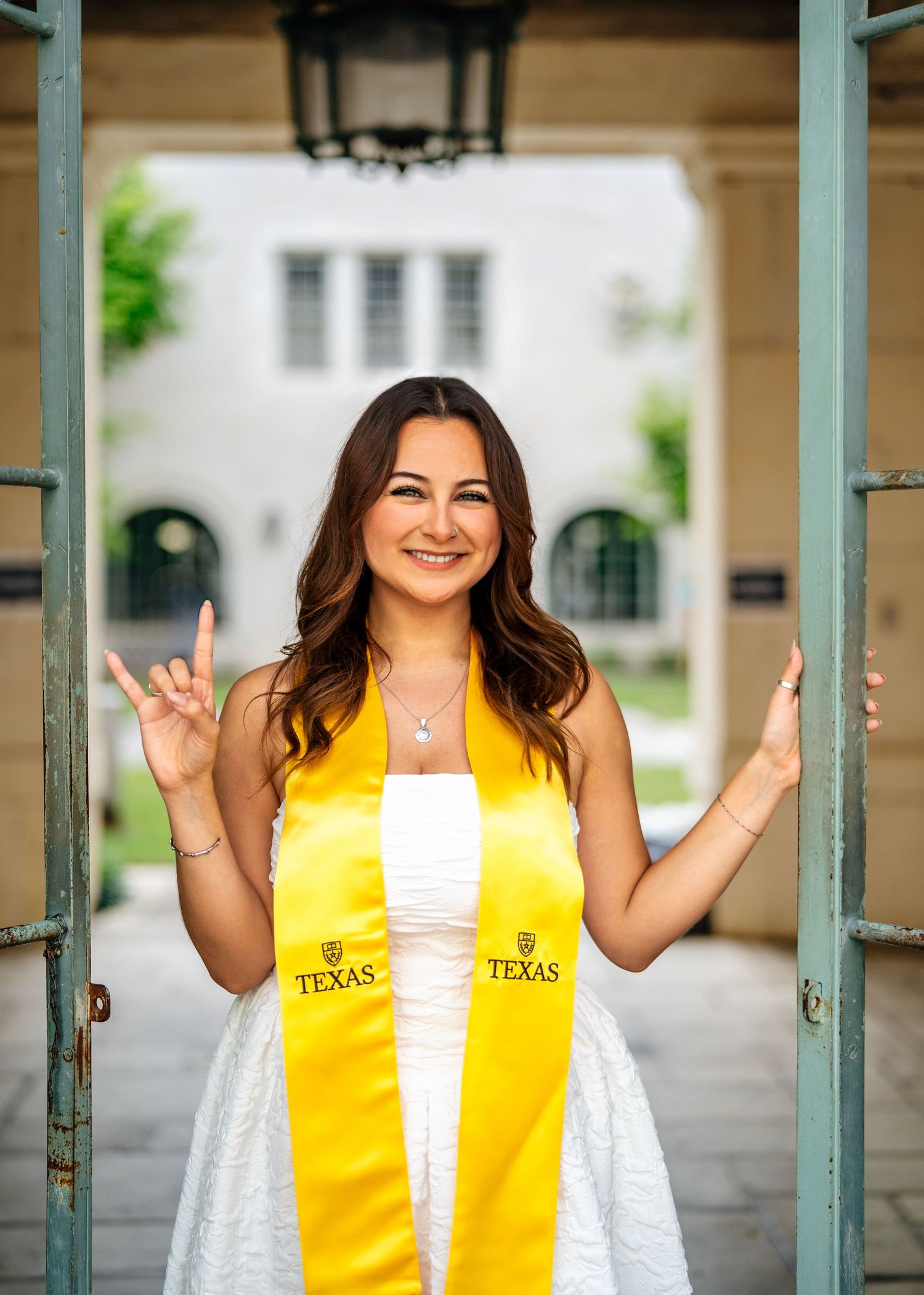 Graduate in bright yellow stole and white graduation gown poses joyfully under outdoor archway.