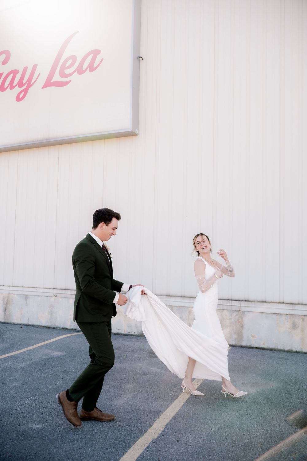 A bride in a flowing white dress and groom in black suit playfully dance together against a white wall.