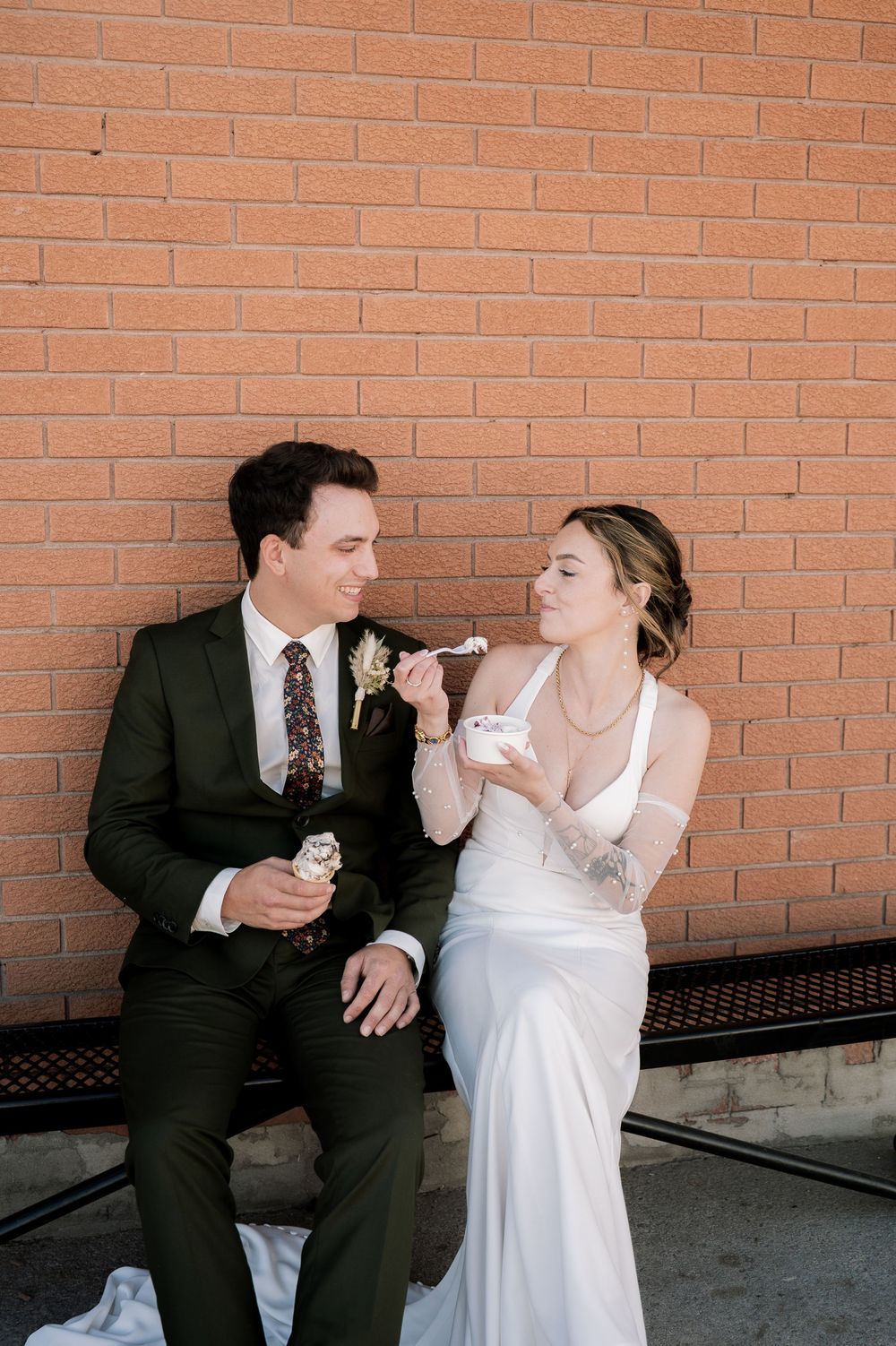 Couple in wedding attire sharing a lighthearted moment against a brick wall background.