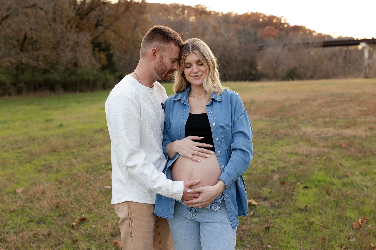 A couple stands together in a grassy field at sunset while lovingly cradling a baby bump.