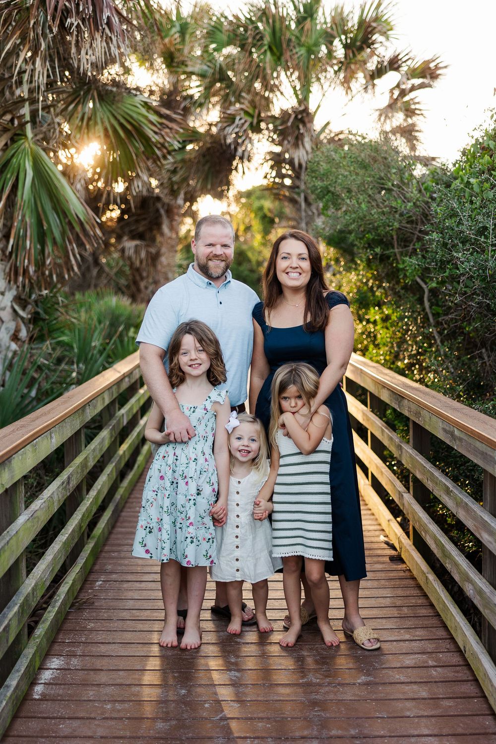 A family poses together on a wooden boardwalk at sunset with palm trees and greenery in the background.