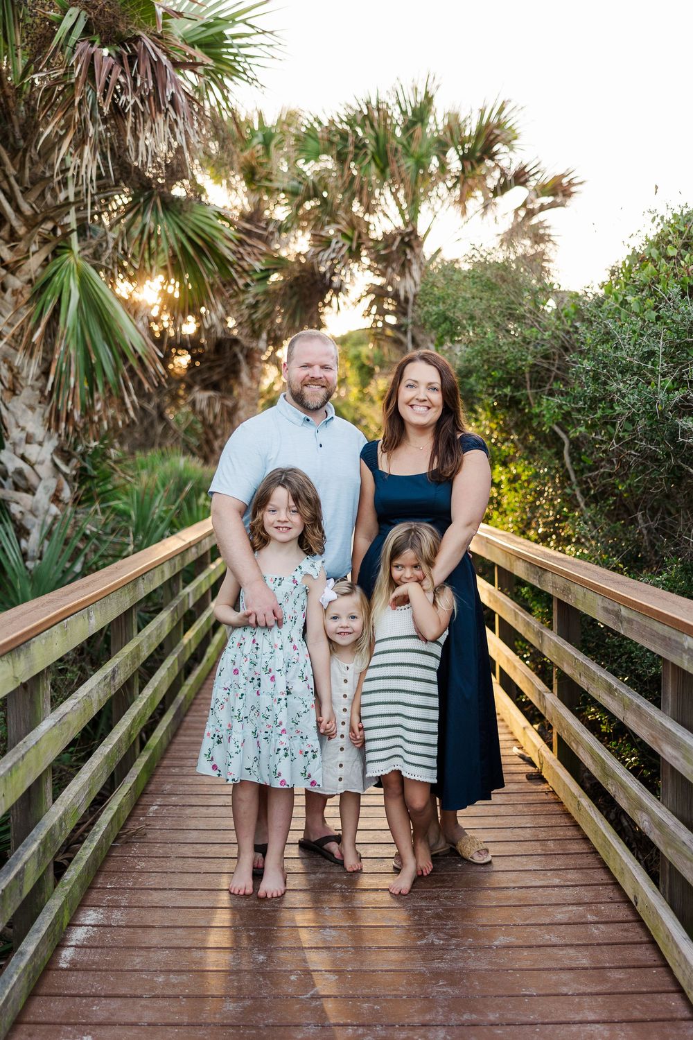 Family laughing together while standing on a wooden boardwalk surrounded by palm trees at sunset.