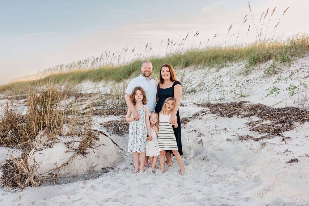 Family group wearing white and navy blue outfits poses together on sandy beach dunes at sunset.