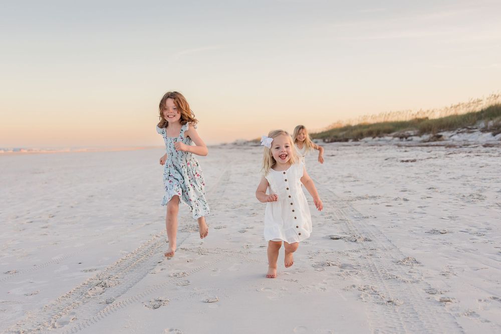 Two children in summer dresses run and play together on a white sandy beach at sunset.