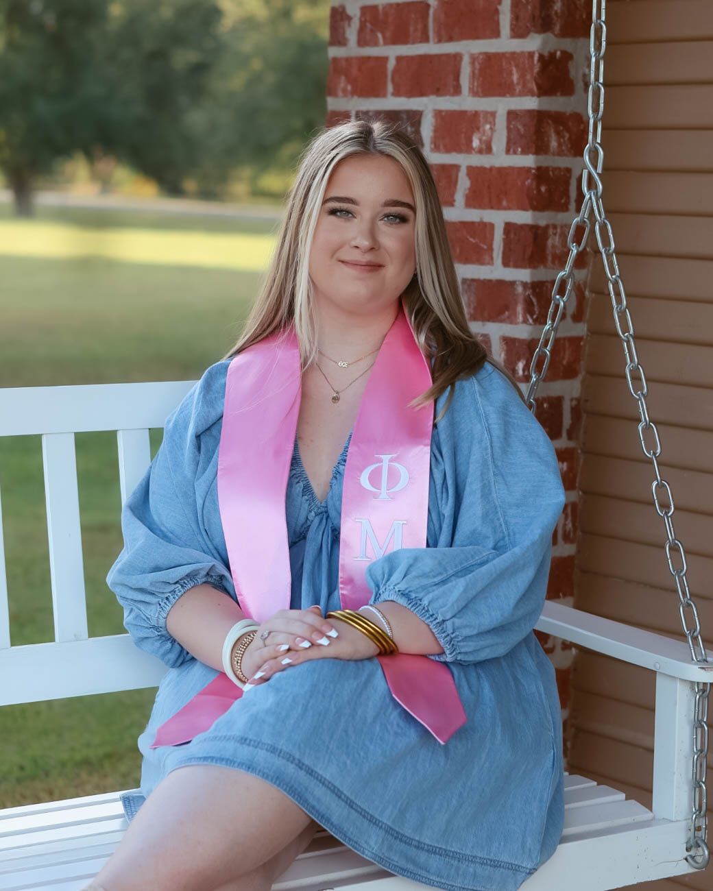 Person in denim jacket with pink sash relaxing on a white porch swing.
