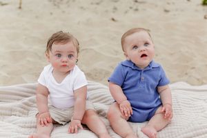 Two babies in casual outfits sitting together on a blanket at the beach.