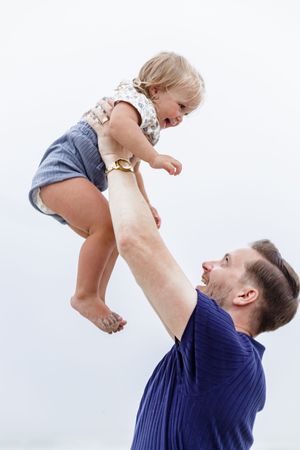 Parent lifting and playing with a joyful toddler against a bright beach background.