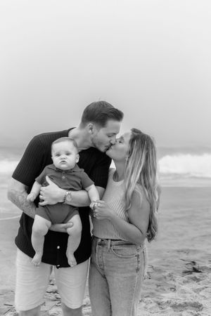 A couple shares tender moments with their baby on a misty beach in a series of black and white photos.