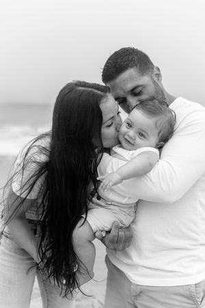 A tender family moment captured in black and white as parents embrace their baby at the beach.